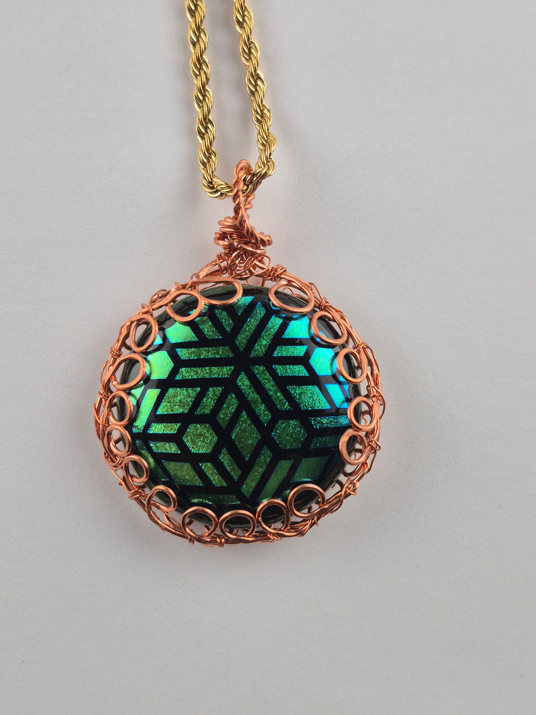A handmade circular pendant necklace featuring a green and black geometric patterned center, wrapped in copper-colored wire, hanging from a gold chain against a neutral gray background.